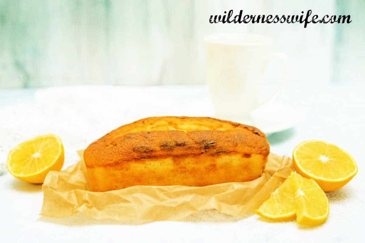 Loaf of KitchenAid Lemon Tea Bread sitting on top of wrinkled parchment paper on a cutting board that is beside to whole lemons.